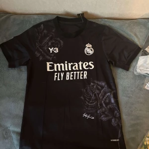 Real Madrid fotbollströja/jersey - Säljer en snygg svart fotbollströja från Y-3 med Real Madrid-logga och 'Emirates Fly Better' tryck på framsidan. Tröjan har korta ärmar och ett coolt ros-mönster. Perfekt för alla fotbollsfans!