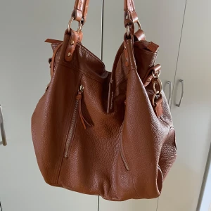 Handväska - Superfin handväska i brunt läder med guld detaljer. Rymmer både dator och anteckningsblock. Den är i väldigt bra skick, utan några slitningar och defekter vad jag kan se.