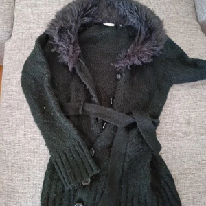 Svart stickad kappa från Lindex - Säljer en svart stickad kappa från Lindex med en fuskpälsdetalj vid kragen. Den har långa ärmar och knäpps med knappar framtill. Dessutom finns ett bälte i midjan 🖤 obs betalning sker via swish 