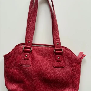 Röd axelväska i nyskick! - Säljer en snygg röd axelväska. Märke Funbag. Väskan är förmodligen i skinn, hittar inte info, och har en praktisk dragkedja upptill. Insidan är fodrad med randigt tyg och har många fack, extra ficka med dragkedja. Hämtas på Hägerstensåsen eller ök. 