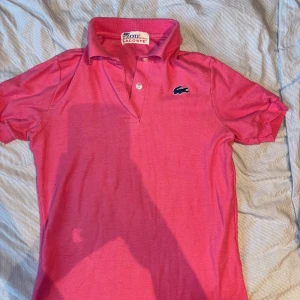 Rosa pikétröja från Lacoste - Säljer en snygg rosa pikétröja från Lacoste. Den har en klassisk krage med knappar och det ikoniska Lacoste-märket på bröstet. Perfekt för en casual look eller en dag på golfbanan. Passar både till jeans och shorts!