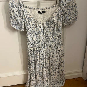 Blommig klänning från Bikbok - Söt blommig klänning från bikbok i vitt och blått. Den har korta puffärmar och en smickrande v-ringad hals med knytning. Perfekt för sommarens alla tillfällen! 🌸
