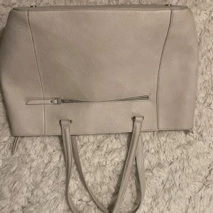 Beige axelväska med dragkedja - Säljer en stilren beige axelväska i syntetmaterial. Väskan har en praktisk dragkedja på framsidan och långa handtag som gör den lätt att bära över axeln. Nypris 800kr.