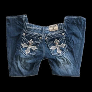 Blå jeans med broderade detaljer - Snygga blå jeans från Live & Fight med unika broderade korsdetaljer på bakfickorna. Jeansen har en cool vintage-look med kontrastsömmar och metallknappar. 