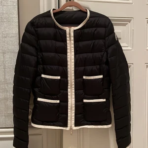 Svart dunjacka från Moncler - Säljer en snygg svart dunjacka från Moncler. Den har en stilren design med vita detaljer och två praktiska fickor fram. Jackan är perfekt för kyliga dagar och har en dragkedja framtill. Passar både till vardags och lite finare tillfällen.