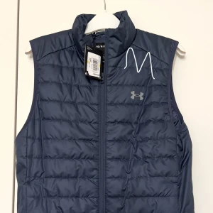 Blå dunväst från Under Armour - Säljer en snygg blå dunväst från Under Armour i storlek M. Västen har en dragkedja framtill och är perfekt för kyliga dagar. Den är quiltad och har en sportig stil, idealisk för både vardag och träning. Passar perfekt till höst och vår! Ny med lappen kvar!