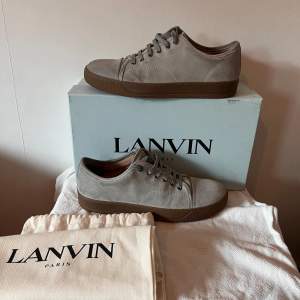 Säljer ett par snygga grå sneakers från Lanvin i mocka. De har en stilren design med snörning och en brun sula som ger en cool kontrast. Perfekta för både vardag och fest! Skorna är i mycket bra skick och kommer med originalkartong samt dustbag och extra skosnören.