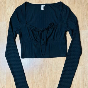Ny svart söt ribbad topp från Nelly Söt knytning fram Bundlerabatt  - Säljer en ny snygg svart ribbad topp från Nelly. Den är croppad och har långa ärmar, perfekt för både vardag och fest. Toppen har fyrkanttig ringning med en söt knytning framtill som ger en extra detalj. Den är i storlek S och sitter tight och bekvämt. 41 cm lång, bystomfång 82 cm, axlar 37 cm, midja 68 cm. Stretchig. bundlerabatt. 
