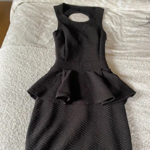 Svart klänning med volangdetaljer - Säljer en elegant svart klänning med en unik öppen rygg och volangdetaljer i midjan. Klänningen har en tight passform och är ärmlös, vilket gör den perfekt för festliga tillfällen. Materialet har en subtil textur som ger en extra touch av stil.