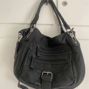 Svart axelväska med dragkedjor och spänne - Säljer en snygg svart axelväska i syntetmaterial. Väskan har flera praktiska dragkedjefickor på framsidan och ett dekorativt spänne. Den har både korta handtag och en justerbar axelrem, vilket gör den perfekt för både vardag och fest. Insidan är rymlig med flera fack för enkel organisering.