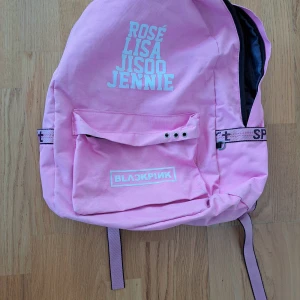 Rosa ryggsäck med Blackpink-tryck - Säljer en superfin rosa ryggsäck med Blackpink-tryck. Den har namnen Rosé, Lisa, Jisoo och Jennie tryckta på framsidan. Ryggsäcken har en praktisk framficka med knappar och justerbara axelremmar med texten 'Sport'. Perfekt för skolan eller utflykter!