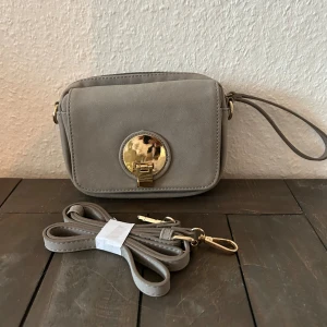 Grå axelväska med gulddetaljer - Säljer en snygg grå axelväska från Pieces. Väskan har en stilren design med en praktisk dragkedja och en justerbar axelrem. Den har också en elegant guldfärgad låsdetalj på framsidan. Nyskick