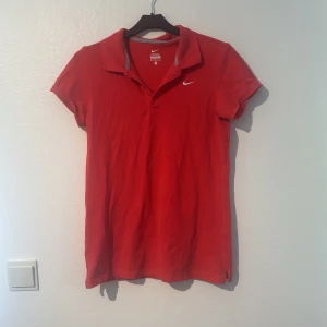 Röd pikétröja från Nike - Säljer en snygg rosa röd pikétröja från Nike i storlek M. Tröjan har korta ärmar och en klassisk krage med knappar. Perfekt för en sportig look eller en avslappnad dag. Den är i ny skick då den aldrig är använd!
