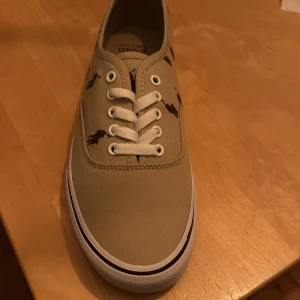 Beige sneakers från Polo Ralph Lauren - Säljer ett par snygga beige sneakers från Polo Ralph Lauren. Skorna har ett coolt mönster med små ponnyer och är tillverkade i bomull. De har en klassisk snörning och en vit sula med märkets logga på baksidan och undersidan. Perfekta för en avslappnad stil!