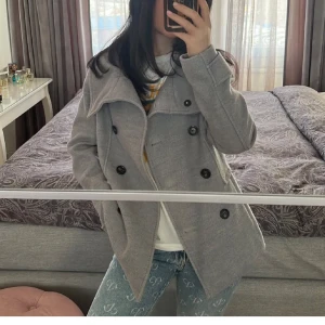 Grå kappa med knappar 💞 - Säljer en snygg grå kappa med stora knappar framtill. Den har en stilren design med en bred krage och är perfekt för höst och vår. Jackan är långärmad och ser ut att vara i bra skick. Perfekt för att hålla sig varm och stilren under kyligare dagar.💞