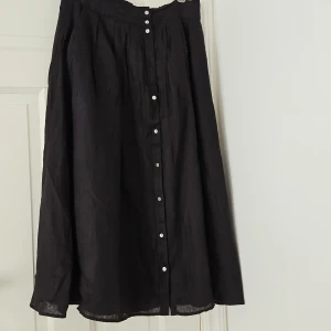 Svart långkjol med knappar - Säljer en elegant svart långkjol med knappar framtill. Kjolen har en klassisk och tidlös design som passar perfekt för både vardag och fest. Den är gjord i linne med underkjol i bomull. Från MQ