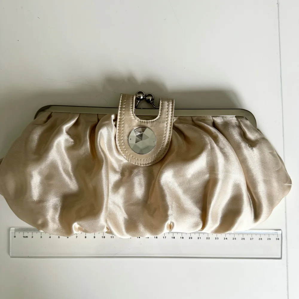 Beige/vit kuvertväska/clutch från Asos. Väskan har en glansig finish och en dekorativ detalj med en stor pärla på. Innerficka med dragkedja. Perfekt för festliga tillfällen! Hämtas på Hägerstensåsen eller ök 🤍. Väskor.