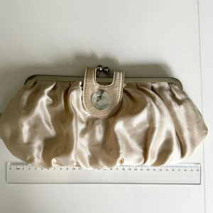 Benvit kuvertväska från Asos - Beige/vit kuvertväska/clutch från Asos. Väskan har en glansig finish och en dekorativ detalj med en stor pärla på. Innerficka med dragkedja. Perfekt för festliga tillfällen! Hämtas på Hägerstensåsen eller ök 🤍