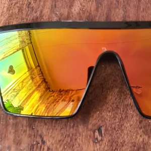 Sportiga solglasögon med spegelglas - Säljer ett par riktigt coola sportiga solglasögon med spegelglas. De har en futuristisk design med en stor, sammanhängande lins i orange och gult som ger en spegeleffekt. Ramen är svart och ger en snygg kontrast till de färgglada glasen. Perfekta för både träning och sommarhäng!
