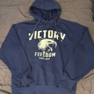 Säljer en blå hoodie med ett coolt tryck av en örn och texten 'Victory Freedom Since 1991'. Den är superbekväm och perfekt för både chill och vardag. Har justerbar huva med snören. Perfekt för höst och vinter!