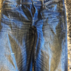 Blå jeans från SMOG - Säljer ett par snygga blå jeans från SMOG. De har en klassisk femficksdesign och en cool tvättad look med lite slitningar. Perfekta för en avslappnad stil. Passar bra till både vardag och fest!