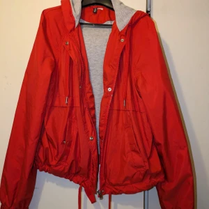 Röd vindjacka med huva - Snygg röd vindjacka med grå huva och dragkedja framtill. Jackan har snörning i midjan och praktiska fickor med knappar. Perfekt för blåsiga dagar.