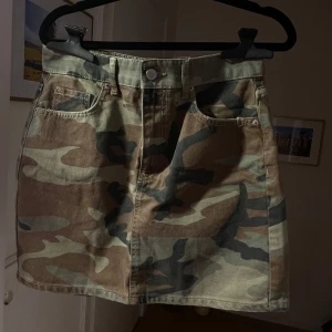 Camouflage kjol - Snygg kortkjol i camouflage-mönster med klassisk jeansstil. Kjolen har fickor fram och bak samt en knapp och dragkedja i midjan. Perfekt för en trendig och avslappnad look.