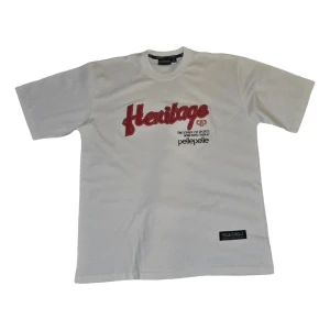 Vit t-shirt från Pelle Pelle - Snygg vit t-shirt från Pelle Pelle med röd broderad text 'Heritage' på framsidan. Perfekt för en casual look med en touch av sportig stil. Rund hals och kortärmad design.