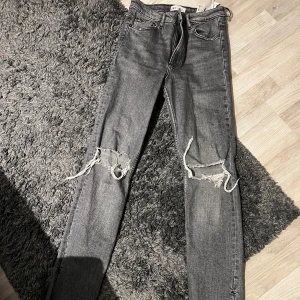 Grå jeans med slitningar från Zara - Snygga grå jeans från Zara med slitningar på knäna. De har en klassisk femficksdesign och dragkedja. Perfekta för en avslappnad stil. De är förkortade där nere så de är även inte överdrivet långa !! Säljs inte längre 