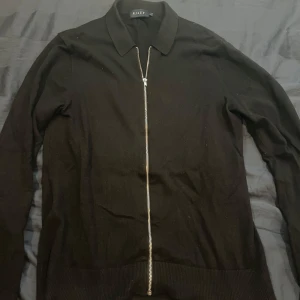 Svart tröja med dragkedja från Riley - Snygg svart tröja från Riley med dragkedja framtill. Tröjan har långa ärmar och en klassisk krage, perfekt för en stilren look. Priset kan diskuteras!