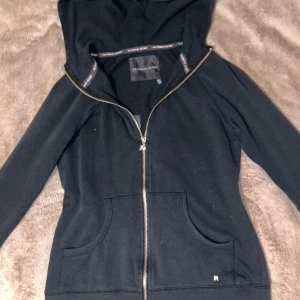 Svart hoodie från Victoria's Secret - Snygg svart hoodie från Victoria's Secret med dragkedja och huva. På baksidan finns texten 'LOVE' i guldiga paljetter och ett par änglavingar. Perfekt för en avslappnad stil med en touch av glamour.