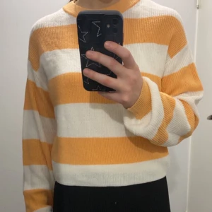 Randig stickad tröja i orange och vit - Säljer en superfin randig stickad tröja i orange och vit. Den är perfekt för höst och vår med sina långa ärmar och mjuka material. Tröjan är croppad och har en normal passform. Perfekt för en avslappnad och stilren look!