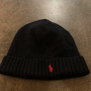 Mörk blå mössa från Polo Ralph Lauren - Snygg mörk blå mössa från Polo Ralph Lauren med röd broderad logga. Perfekt för kyliga dagar med sin ribbade kant som ger extra värme och stil. Mössan är också mörkblå men bilden gör så den ser väldigt svart ut.