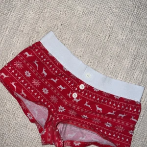 Söta pyjamashorts  - Säljer ett par röda shorts med julmönster i vitt, inklusive renar och snöflingor. De har en bekväm midja och knappar framtill. Perfekta för mysiga vinterdagar!