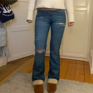 Blå bootcut jeans med slitningar - Snygga blå bootcut jeans från Wrangler med slitningar på låren och knäna. De har en klassisk femficksdesign och en läderpatch med logotyp på bakfickan. Lågmidjade!! W26