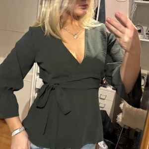 Mörkgrön omlottopp - Snygg mörkgrön omlottopp med v-ringning och knytband i midjan. Toppen har trekvartsärmar med lätt volangdetalj vid ärmsluten. Perfekt för en stilren look.