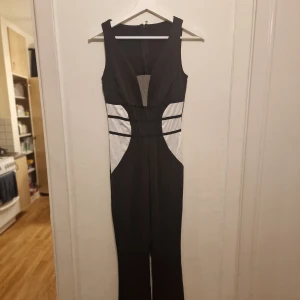 Svart och vit jumpsuit - Snygg svart och vit ärmlös jumpsuit med v-ringning och dragkedja i ryggen. Den har kontrasterande vita paneler i midjan som ger en smickrande siluett. Perfekt för en stilren look. Garderobsrensning så kika gärna på övriga auktioner.