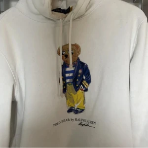 Vit hoodie med Polo Bear från Ralph Lauren - Säljer en vit hoodie från Ralph Lauren med det ikoniska Polo Bear-trycket på framsidan. Tröjan har en justerbar huva med snören och långa ärmar. Perfekt för en avslappnad stil.