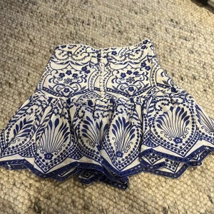 Vit och blå kjol med mönster - Charmig kortkjol i vitt med blått mönster. Kjolen har en volangdetalj och knappar framtill, vilket ger en söt och lekfull stil. Perfekt för varma dagar.