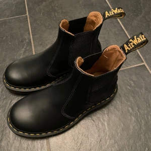Svarta Dr Martens - Snygga svarta Chelsea boots i skinn från AirWair med klassisk gul söm runt sulan. De har elastiska paneler på sidorna och en praktisk dragflik baktill för enkel påtagning.