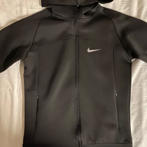 Nike Hoodie - Snygg svart hoodie från Nike med dragkedja och huva. Perfekt för en sportig look eller avslappnade dagar. Den har en diskret Nike-logga på bröstet och långa ärmar.
