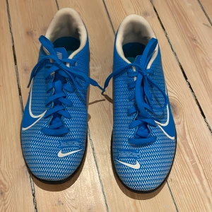 Blå Nike Mercurial inomhus fotbollsskor - Säljer ett par blå Nike Mercurial inomhus fotbollsskor med vit logga och snörning. Skorna har en mönstrad ovandel och svart sula med texten 'JUST DO IT' på sidan. Perfekta för många olika aktiviteter!