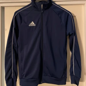 Mörkblå träningsjacka från Adidas - Säljer en mörkblå träningsjacka från Adidas med dragkedja och vita detaljer. Jackan har en hög krage och långa ärmar, perfekt för sportiga aktiviteter. Adidas-loggan är tryckt på bröstet.