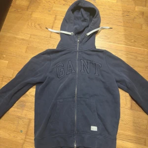 Mörkblå hoodie från GANT - Säljer en snygg mörkblå hoodie från GANT med dragkedja och stor logga på bröstet. Tröjan har en justerbar huva med vita snören och praktiska fickor framtill. Perfekt för en avslappnad stil.