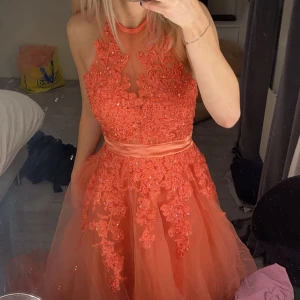 Orange spetsklänning - Fantastisk orange långklänning med spetsdetaljer och broderier. Klänningen har en öppen rygg och är ärmlös, vilket ger en elegant look. Perfekt för speciella tillfällen.