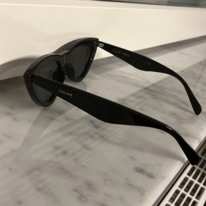 Svarta solglasögon från Celine - Snygga svarta solglasögon från Celine med en elegant cat-eye design. Glasögonen har en stilren svart färg och breda skalmar med Celine-logotypen i guld. Perfekta för att ge en touch av lyx till din look.