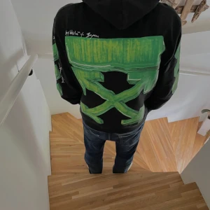 Svart hoodie från Off-White med gröna detaljer - Säljer en svart hoodie från Off-White med coola gröna grafiska detaljer på ryggen och ärmarna. Den har en klassisk känguruficka och justerbar huva. Perfekt för en avslappnad och stilren look!