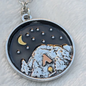 Halsband med nattligt landskap - halsband med ett runt hänge som visar ett nattligt landskap. Hänget har en svart bakgrund med en gul måne och stjärnor. Det finns också ett berglandskap med snötäckta träd och ett tält i mitten. Kedjan är i silverfärgat Metall.