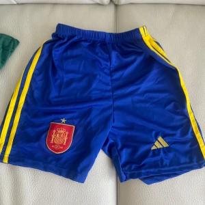 Fotbolls sett Spanien  - Säljer ett par blå fotbollsshorts från Adidas med gula ränder längs sidorna. Shortsen har en elastisk midja och ett tryckt emblem på ena benet. Perfekta för fotbollsträningen eller matchen! Har aldrig används säljer för att har två likadana (Lamine yamal)