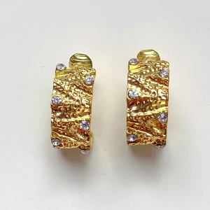 Guldörhängen med glittriga stenar - Säljer ett par snygga guldörhängen med en cool textur och glittriga stenar. Okänd metall. De har en unik design med ett vågigt mönster och är perfekta för att ge lite extra bling till din outfit. Örhängena är i nyskick och har en bekväm clips för enkel användning. Perfekta för fest eller en utekväll!